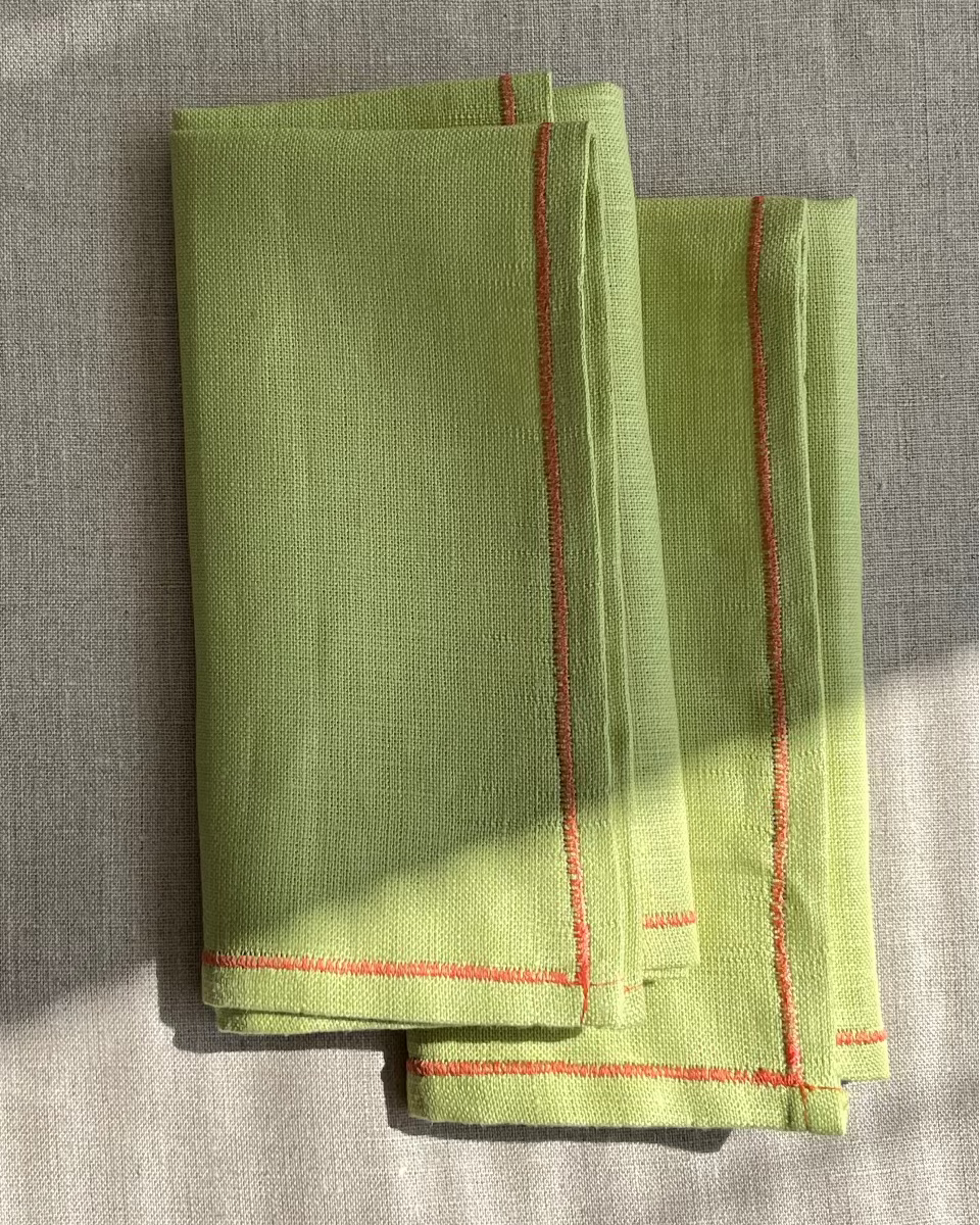 A folded light green linen napkin with red stitched borders, displayed on a textured beige linen surface with natural dappled sunlight.