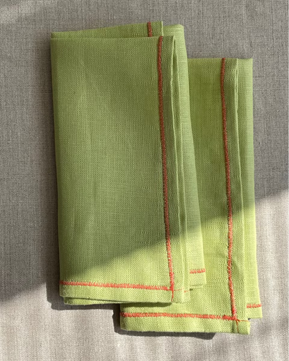 A folded light green linen napkin with red stitched borders, displayed on a textured beige linen surface with natural dappled sunlight.