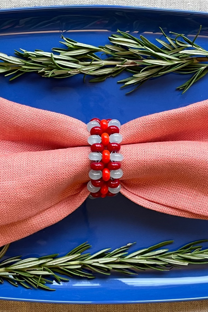 A close-up of a coral pink cloth napkin tied in a bow on a blue plate, secured with a red and white beaded napkin ring, with rosemary sprigs around it.
