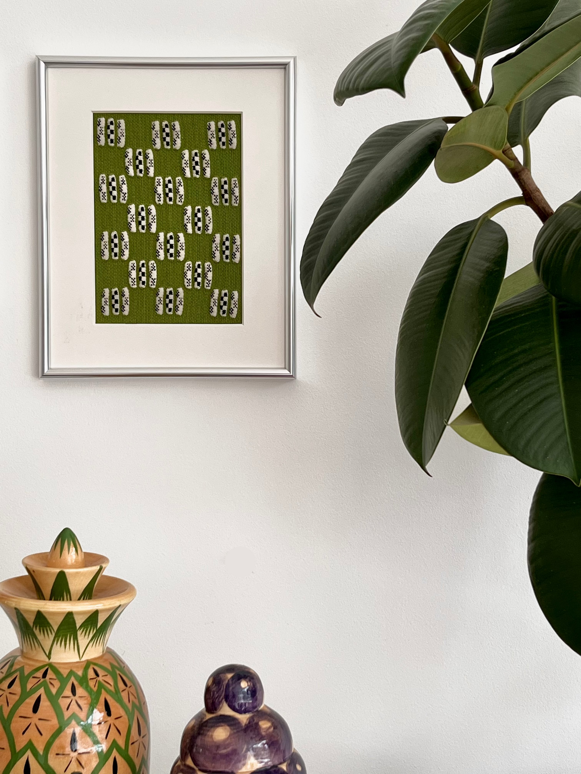 Framed artwork with a green pattern on a white wall next to decorative vases and a plant.