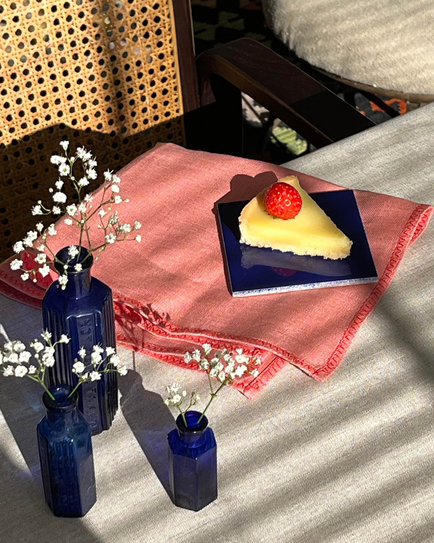 A styled tabletop scene with a slice of yellow cake on a dark blue plate, set on a pink cloth over a striped beige table runner. Three cobalt blue glass vases with small white flowers are arranged nearby, with a woven cane screen and chair visible in the background.