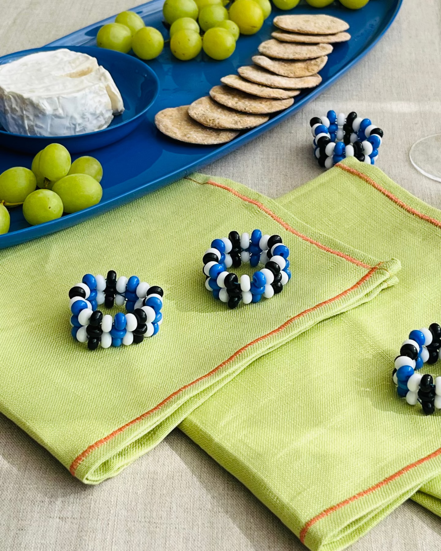 Blue and white beaded bracelet cuffs displayed on folded light green napkins, with a blue charcuterie board holding grapes, crackers, and cheese in the background.