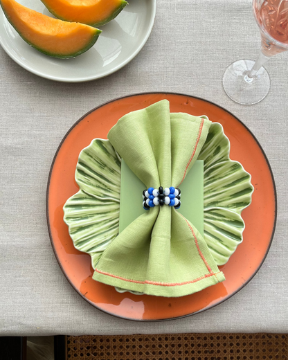 A styled table setting featuring a light green pleated napkin folded into a bow and secured with a blue and white beaded ring, placed on an orange plate. In the background, a white plate holds slices of cantaloupe melon, and a glass of rosé wine is partially visible on a textured beige tablecloth.