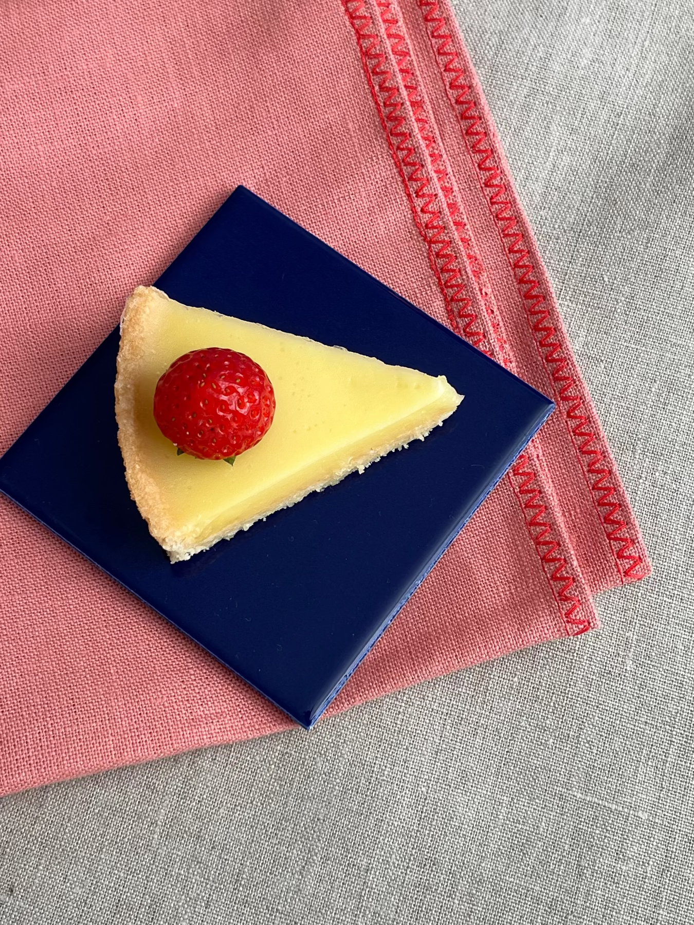 A triangular slice of lemon tart with a whole strawberry on top, presented on a dark blue square plate over a pink napkin on a gray linen surface.