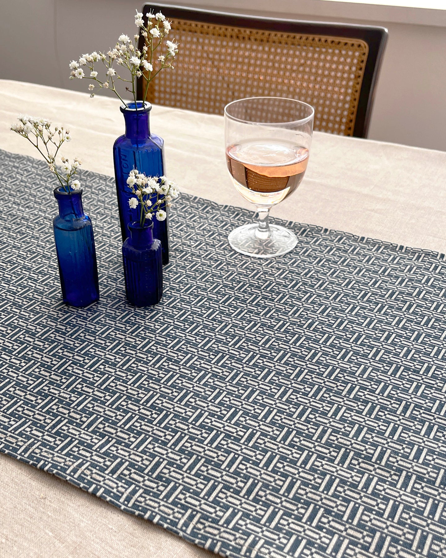 A woven table runner with a geometric basketweave pattern in gray and white, styled on a dining table with blue glass bottles, baby's breath flowers, and a glass of rosé wine.