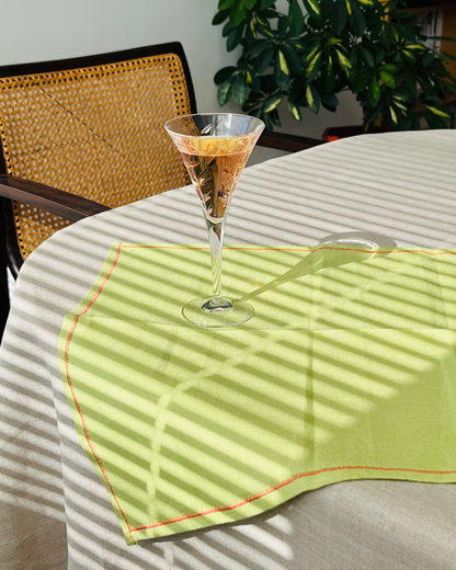 A light green table runner with thin orange-red stitched borders is draped over a round table in a real dining setting. A martini glass with a cocktail sits on top, and strong diagonal window shadows are cast across the runner and table.