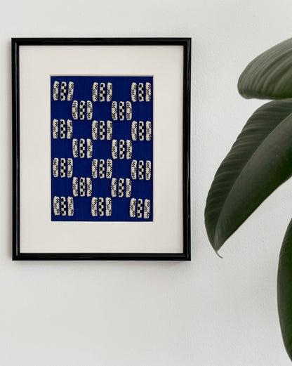 Framed artwork with a blue and white pattern on a white wall with a plant.