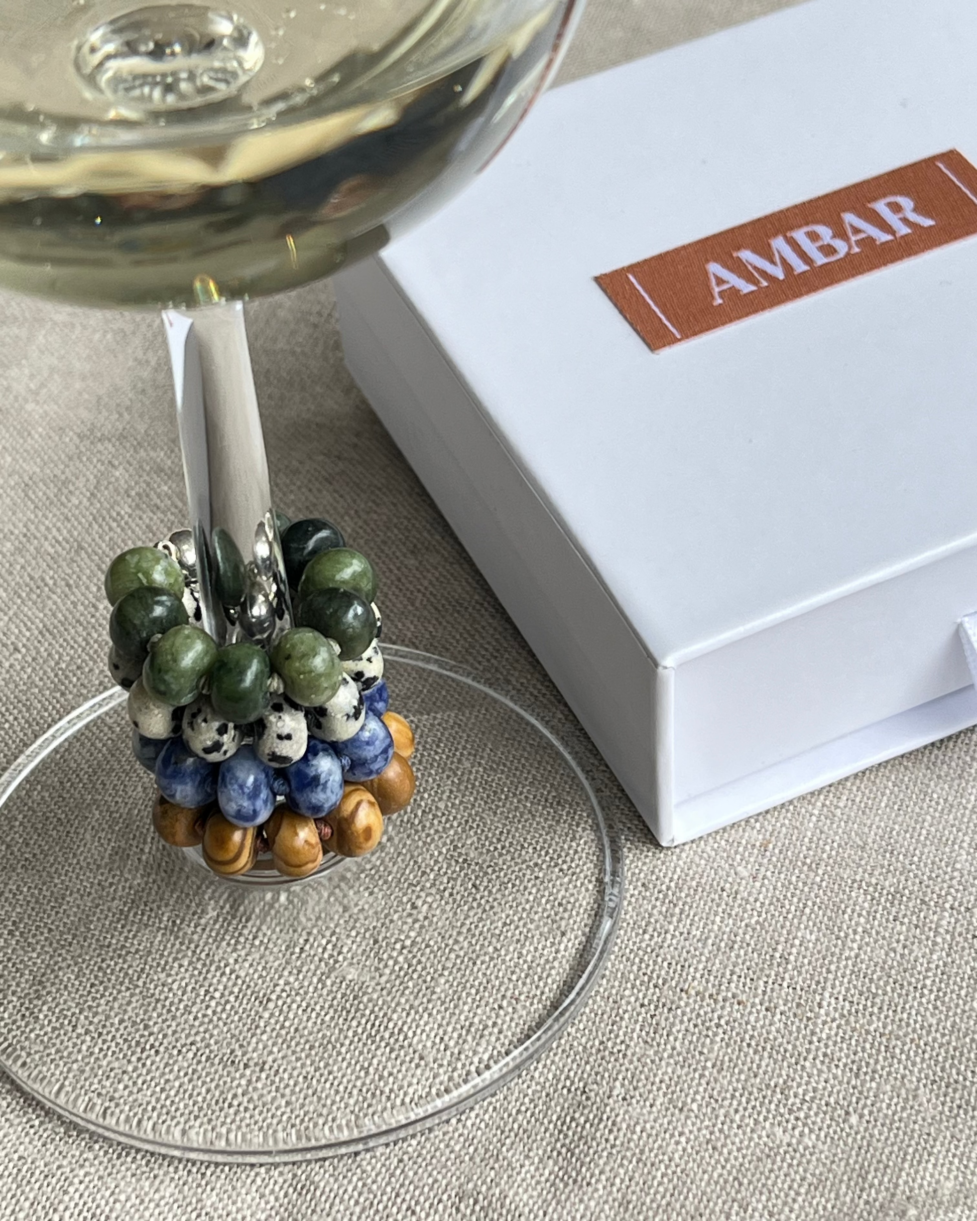 Decorative wine glass charm with colored beads on a glass, next to an 'AMBAR' branded box.