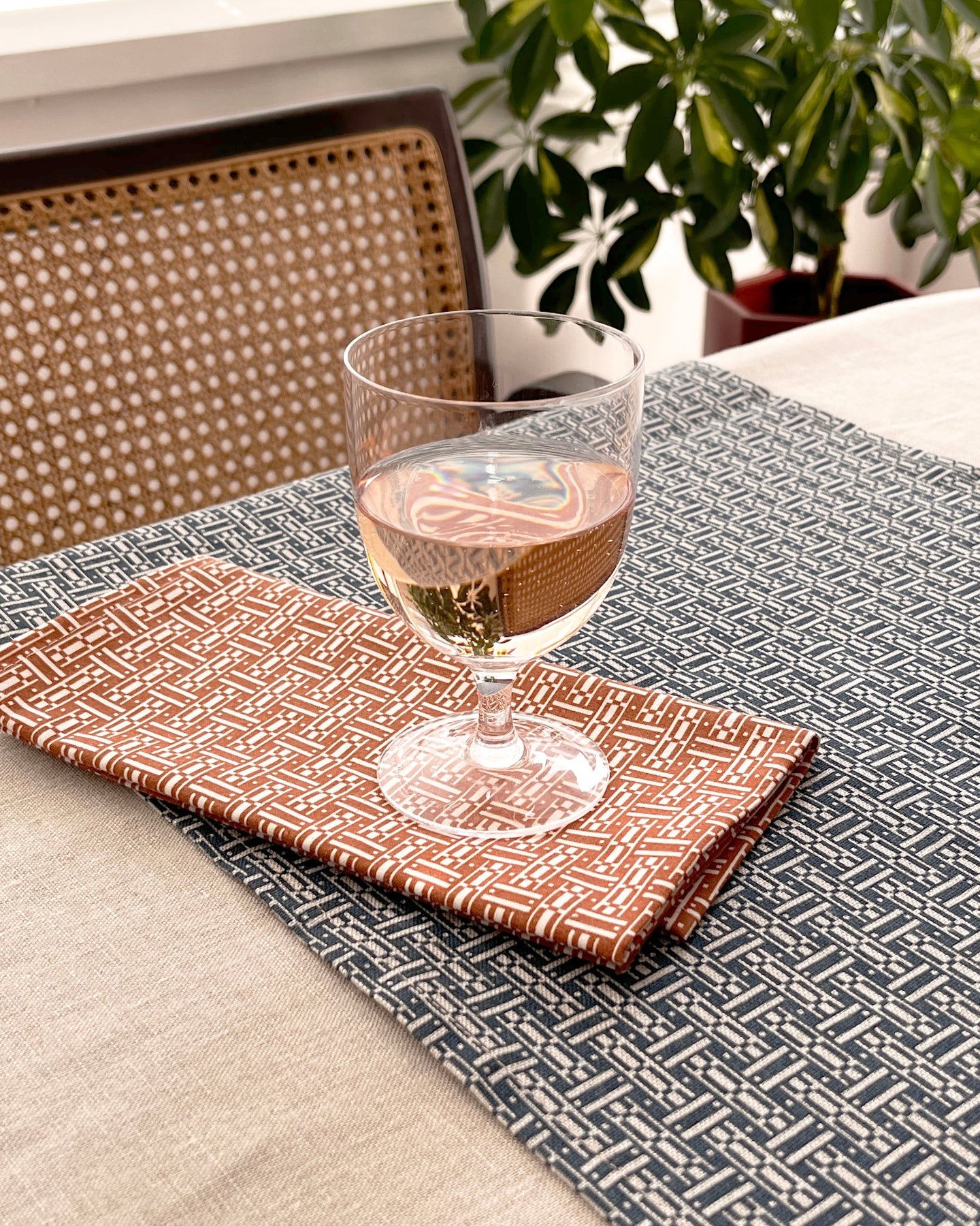A styled dining table setting featuring a dark gray and white geometric patterned runner, a rust-orange patterned napkin, and a clear wine glass filled with white wine, with a woven chair and green plant in the background.