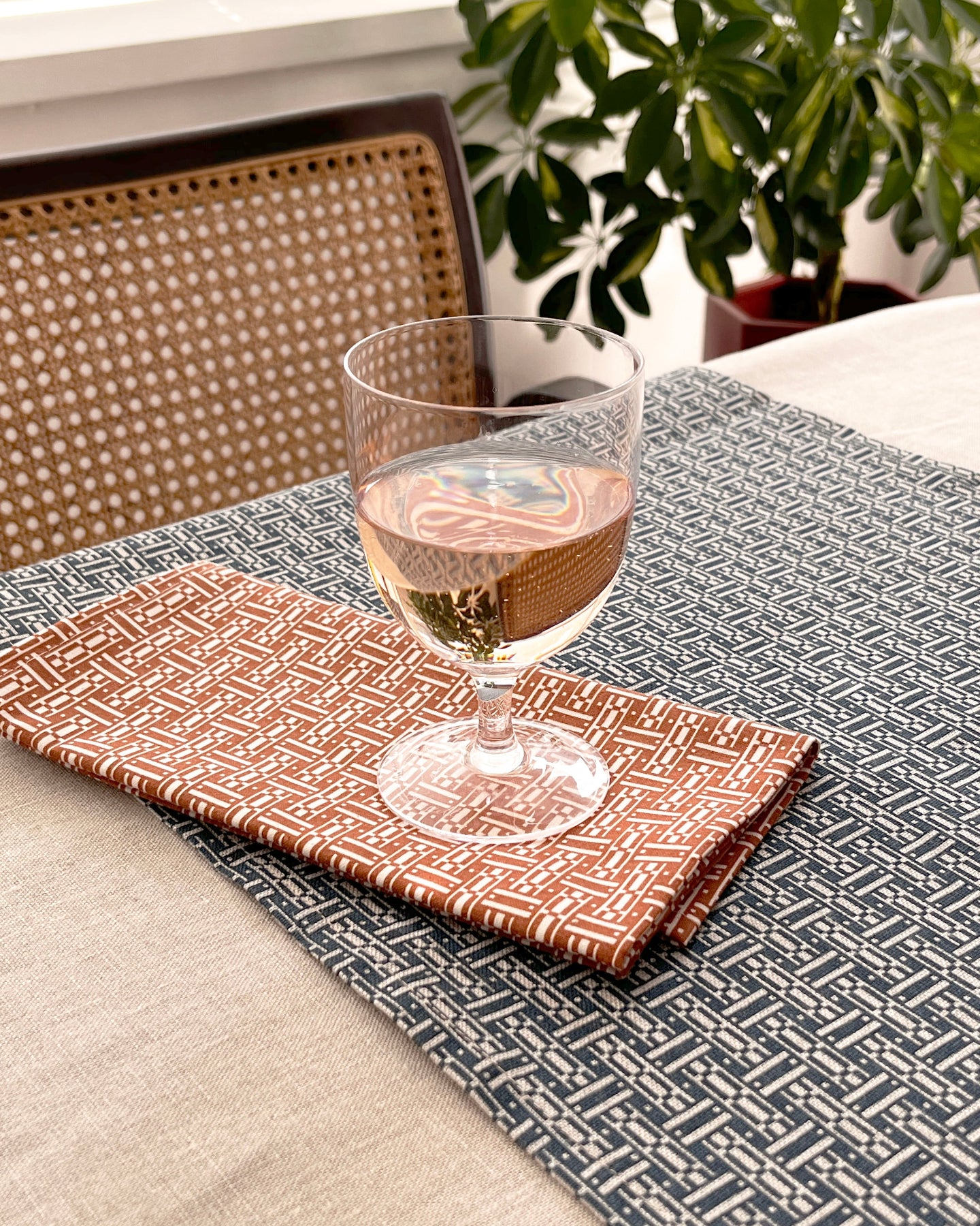 A styled dining table setting featuring a dark gray and white geometric patterned runner, a rust-orange patterned napkin, and a clear wine glass filled with white wine, with a woven chair and green plant in the background.