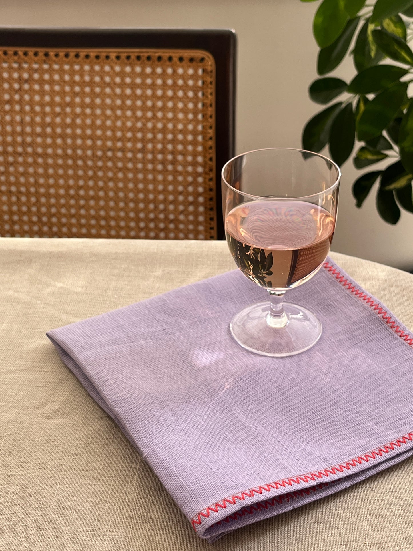 A folded light purple linen napkin with red zigzag stitching, displayed on a beige tablecloth next to a clear wine glass, with a cane-back chair and green leaves in the background.