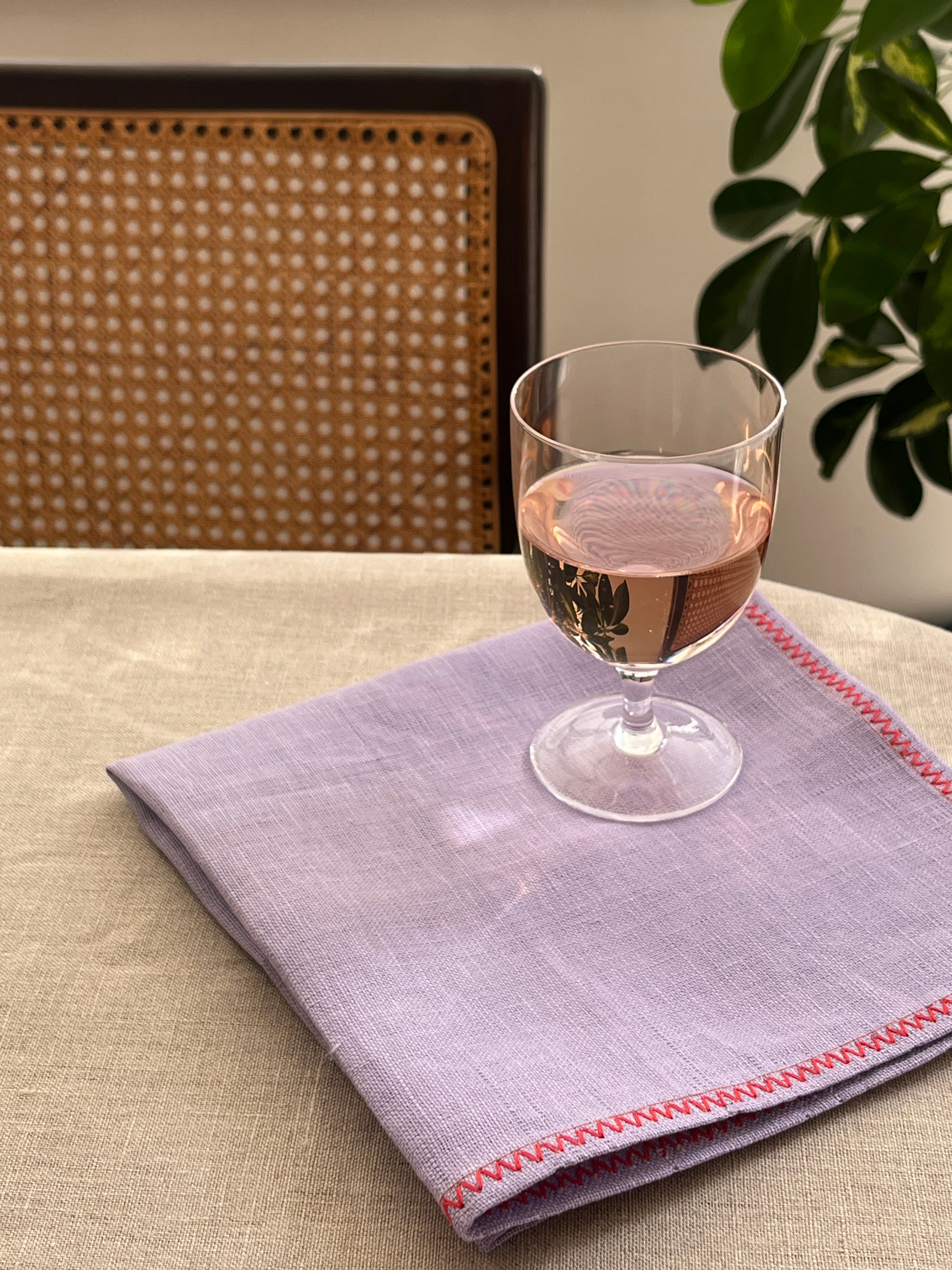 A folded light purple linen napkin with red zigzag stitching, displayed on a beige tablecloth next to a clear wine glass, with a cane-back chair and green leaves in the background.