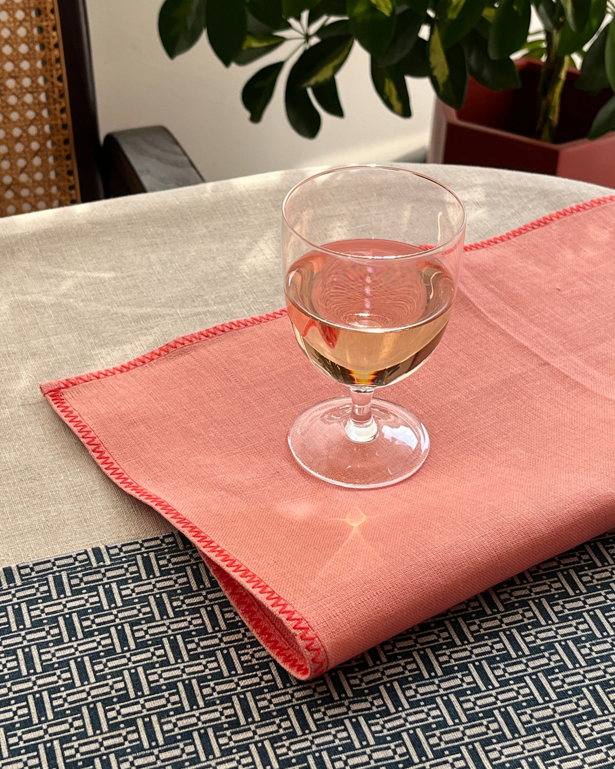 A folded dusty pink linen napkin with red stitched edges, styled on a table alongside a wine glass and a patterned blue and white runner.